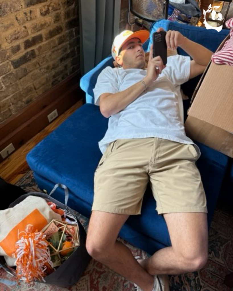 Young man in an orange ball cap lying on a blue futon and scrolling his phone during move in day
