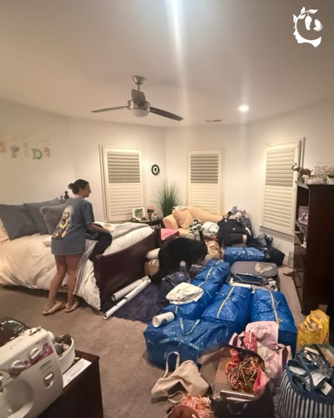 Young woman packing a bedroom filled with blue tote bags and suitcases, with the family dog investigating