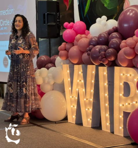 A woman in a floral dress speaking on stage at a WIP (Women in Psychiatry/Professional) event, standing beside large illuminated marquee letters "WIP" surrounded by a pink, mauve, and white balloon arch.
