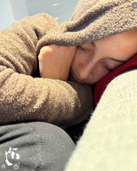 A young person with a small nose ring sleeping peacefully, bundled in a cozy brown fleece hoodie, resting their head on their arms against a red surface.
