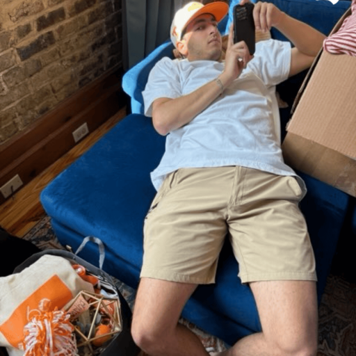 Young man in an orange ball cap lying on a blue futon and scrolling his phone during move in day