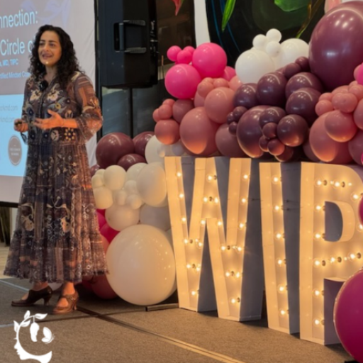 A woman in a floral dress speaking on stage at a WIP (Women in Psychiatry/Professional) event, standing beside large illuminated marquee letters "WIP" surrounded by a pink, mauve, and white balloon arch.