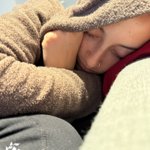 A young person with a small nose ring sleeping peacefully, bundled in a cozy brown fleece hoodie, resting their head on their arms against a red surface.